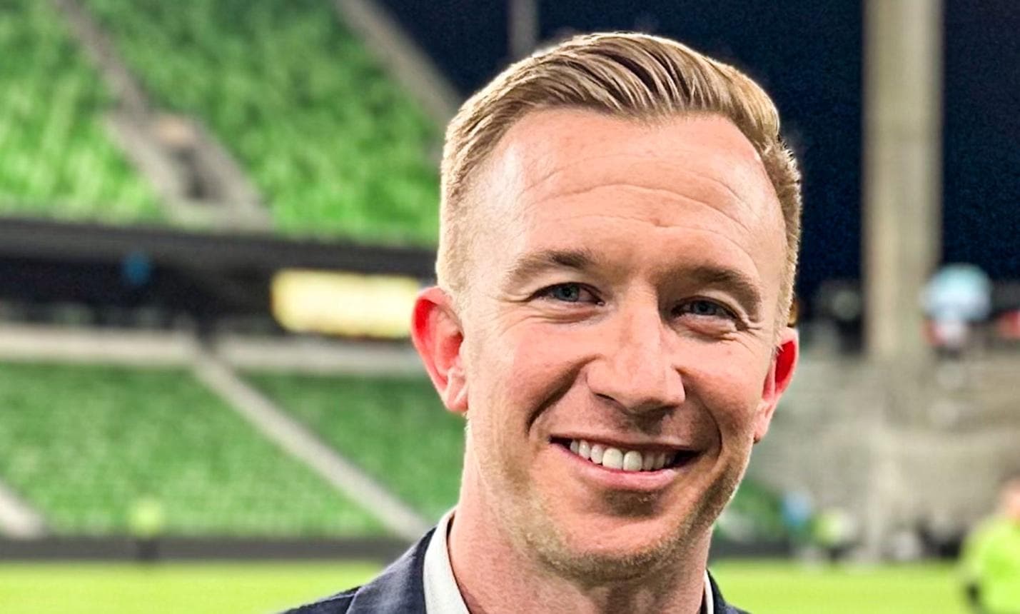 Smiling man with short blonde hair in a dark jacket at a blurred green stadium.
