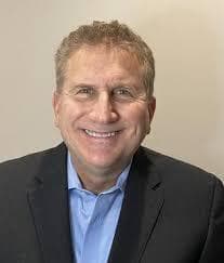Smiling middle-aged man with short hair wearing a blue shirt and a dark blazer.