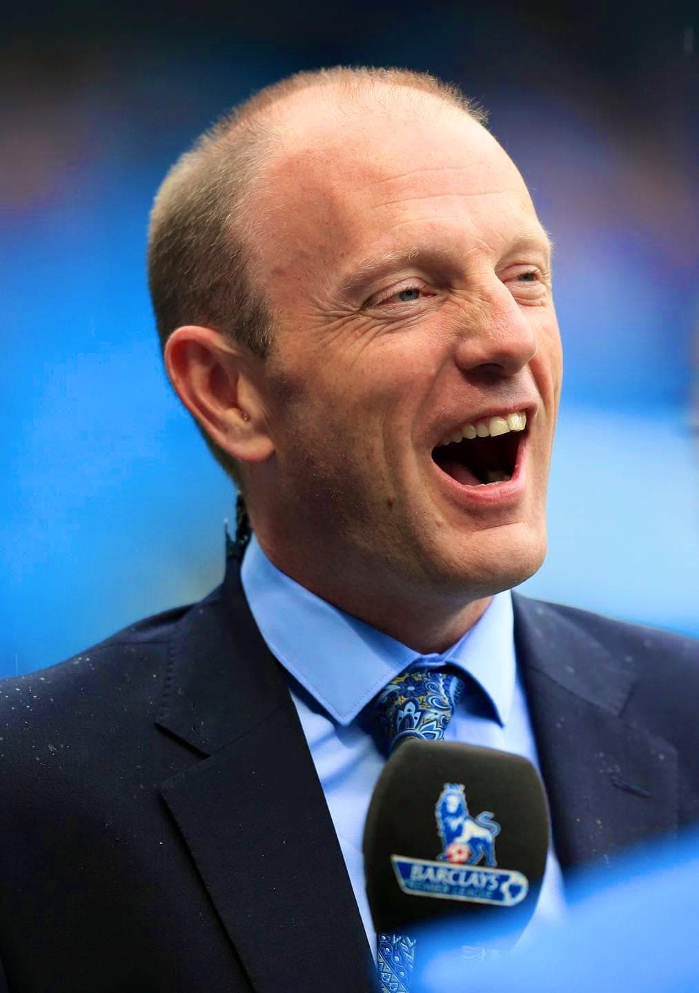 Close-up of a laughing man in a suit behind a Barclays Premier League microphone.