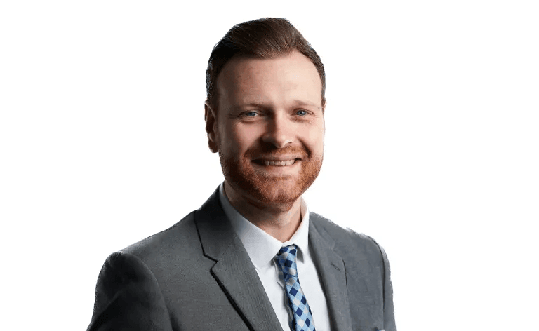 Smiling man with a red beard wearing a grey suit and blue checkered tie.