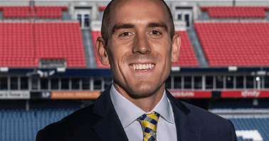 Smiling man in a suit and yellow patterned tie with a large sports stadium background.